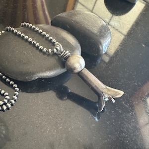 May include: A silver necklace with a unique pendant shaped like a stylized bird with a round ball at the top. The pendant is made of metal and has a textured finish.