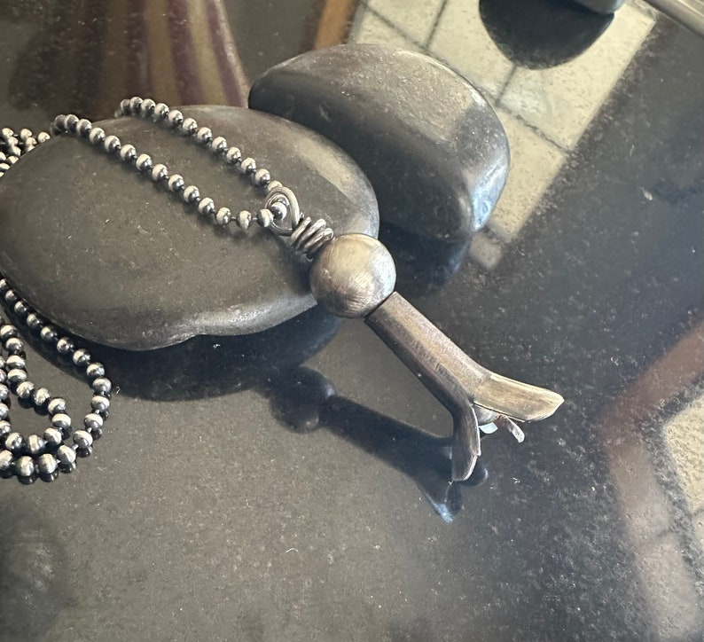 May include: A silver necklace with a unique pendant shaped like a stylized bird with a round ball at the top. The pendant is made of metal and has a textured finish.