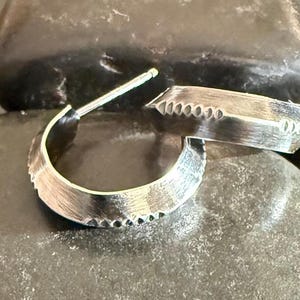 May include: A pair of silver hoop earrings with a textured, hammered finish. The earrings have a unique, geometric design with a pointed edge.