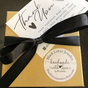 May include: A small, gold-colored box tied with a black satin ribbon. A white card with the words "Thank You" and a heart is on top. A round sticker reads "Black Lotus Jewelry handmade with Love".
