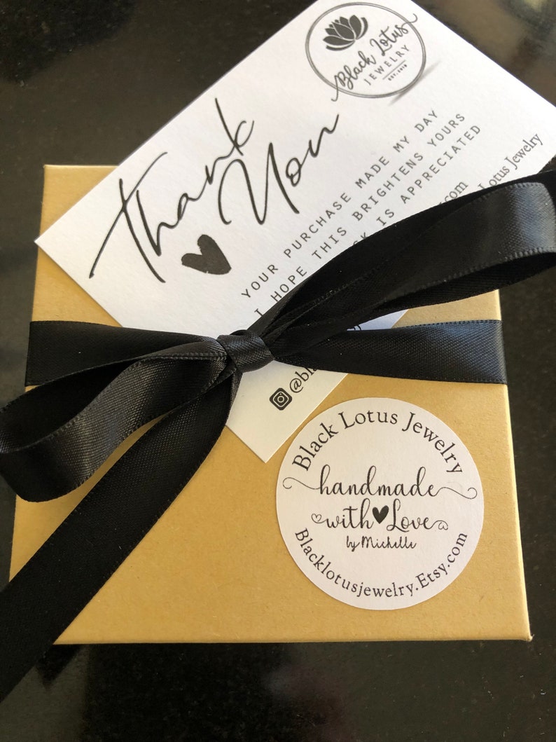 May include: A small, gold-colored box tied with a black satin ribbon. A white card with the words "Thank You" and a heart is on top. A round sticker reads "Black Lotus Jewelry handmade with Love".
