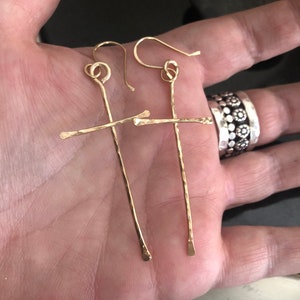 May include: A pair of gold-colored earrings with a cross design. The earrings have a hammered texture and a simple, elegant style. The hooks are curved and the earrings are held in a hand.
