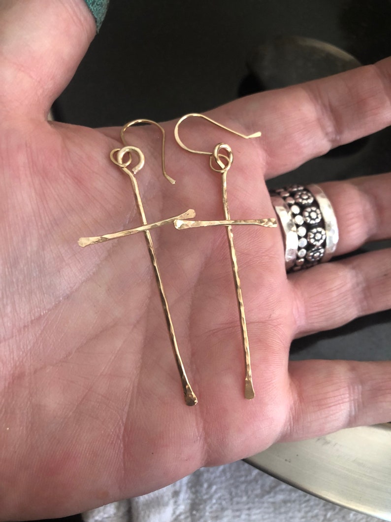 May include: A pair of gold-colored earrings with a cross design. The earrings have a hammered texture and a simple, elegant style. The hooks are curved and the earrings are held in a hand.