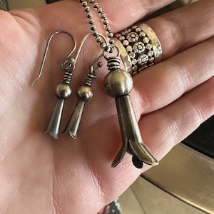 May include: A silver necklace with a pendant shaped like a flower. The pendant is attached to a silver chain. The necklace is also accompanied by a pair of silver earrings with flower-shaped pendants.