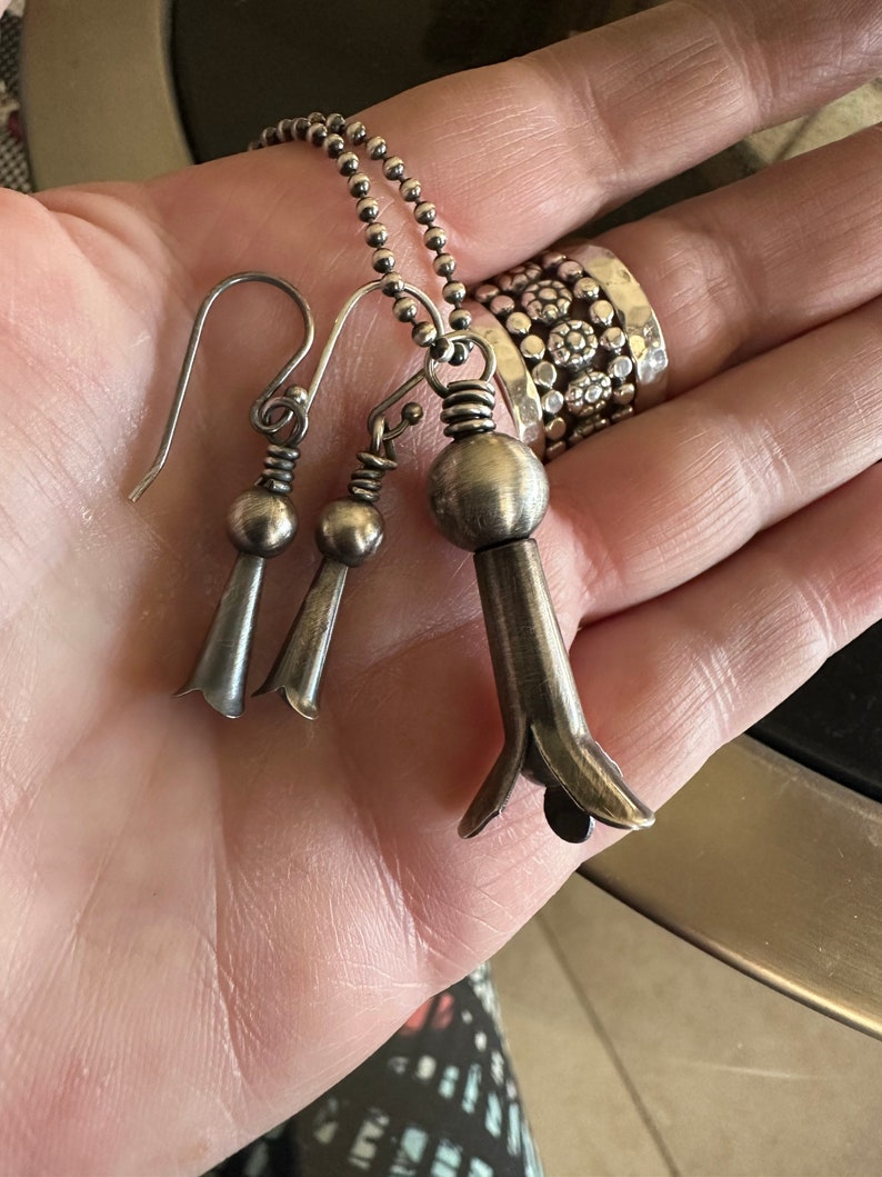 May include: A silver necklace with a pendant shaped like a flower. The pendant is attached to a silver chain. The necklace is also accompanied by a pair of silver earrings with flower-shaped pendants.