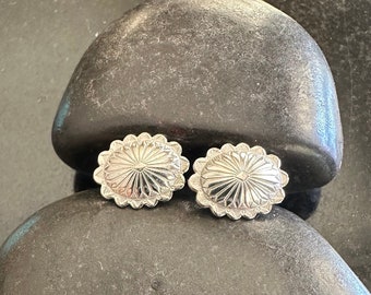 May include: A pair of silver-colored, oval-shaped stud earrings. Each earring features a sunburst design with a scalloped edge. The earrings are displayed on a dark gray, textured surface.