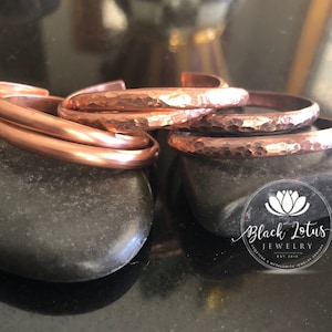 May include: Copper bracelets in various styles, including smooth and hammered textures, are displayed on dark gray stones. The bracelets have a warm, reddish-brown hue, reflecting the light. The jewelry is from Black Lotus Jewelry.