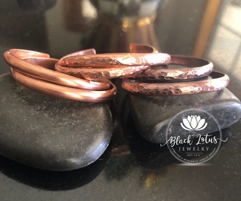 May include: Copper bracelets in various styles, including smooth and hammered textures, are displayed on dark gray stones. The bracelets have a warm, reddish-brown hue, reflecting the light. The jewelry is from Black Lotus Jewelry.