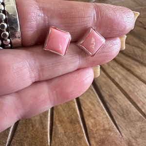 Peut inclure: Une paire de boucles d'oreilles carrées roses avec une bordure argentée. Les boucles d'oreilles sont présentées sur une surface en bois. Elles ont une finition brillante et un design géométrique simple.