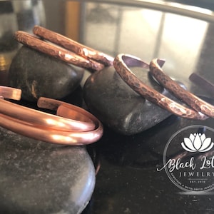May include: Copper bracelets in various styles, including hammered and smooth finishes, are displayed on dark gray stones. The bracelets are a warm, reddish-brown color, reflecting light. The Black Lotus Jewelry logo is visible in the bottom right corner.