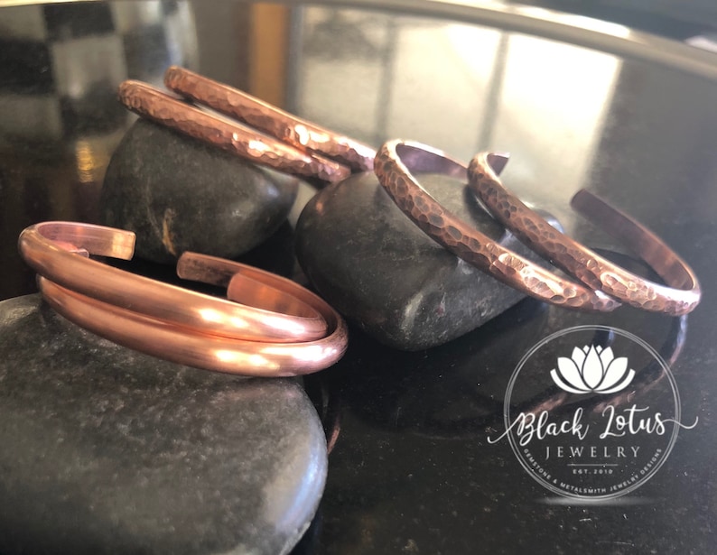 May include: Copper bracelets in various styles, including hammered and smooth finishes, are displayed on dark gray stones. The bracelets are a warm, reddish-brown color, reflecting light. The Black Lotus Jewelry logo is visible in the bottom right corner.