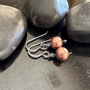 Op de afbeelding: Een paar zilveren oorhangers met koperkleurige ronde kralen. De oorhangers zijn gefotografeerd op een zwart oppervlak met drie gladde grijze stenen.