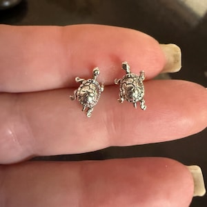 May include: Two small, silver-colored turtle stud earrings. The earrings are detailed with textured shells, tiny legs, and small heads. The earrings are shown on a person's finger.