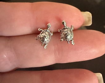 May include: Two small, silver-colored turtle stud earrings. The earrings are detailed with textured shells, tiny legs, and small heads. The earrings are shown on a person's finger.