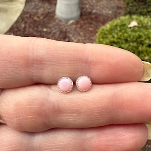 May include: A pair of small, round, pink stud earrings with a silver setting. The earrings are shown on a person's finger.