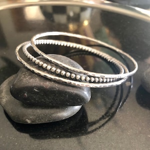 May include: Three silver bangles, one with a hammered finish and two with beaded edges, stacked on top of each other.