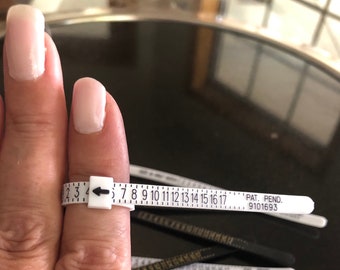 May include: White plastic ring sizer with black markings from 2 to 17. The sizer is wrapped around a finger and the arrow points to size 4. The text "PAT. PEND. 9101693" is printed on the sizer.
