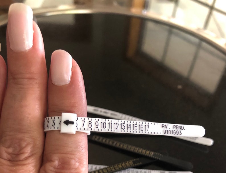 May include: White plastic ring sizer with black markings from 2 to 17. The sizer is wrapped around a finger and the arrow points to size 4. The text "PAT. PEND. 9101693" is printed on the sizer.