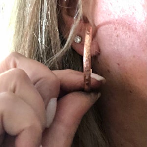 May include: Copper-colored hoop earrings with a hammered texture. The earrings are round and have a simple, elegant design. The image shows a close-up of the earrings being held up to an ear.