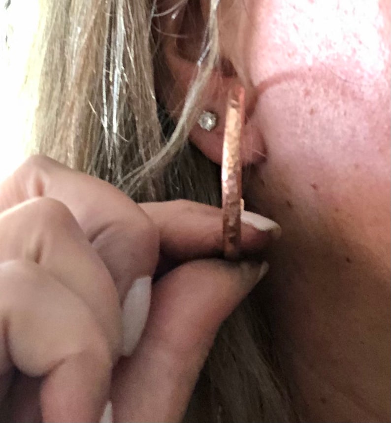 May include: Copper-colored hoop earrings with a hammered texture. The earrings are round and have a simple, elegant design. The image shows a close-up of the earrings being held up to an ear.