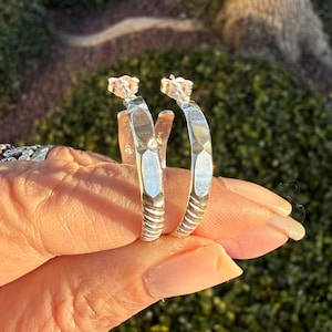 May include: A pair of silver hoop earrings with a textured, hammered design. The earrings have a post backing and are held up by a hand. The earrings are in focus, with a blurred green background.