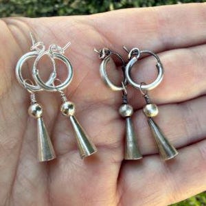 May include: Two pairs of hoop earrings featuring a small silver-toned sphere and a dangling conical charm. The left pair has a bright, polished silver finish, while the right pair displays a darker, oxidized silver-toned metal appearance.