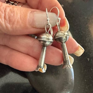 May include: A pair of silver earrings with a distinctive floral design. Each earring has a round top and a bell-shaped bottom. The earrings are suspended by a silver hook. The earrings are displayed on a dark surface.