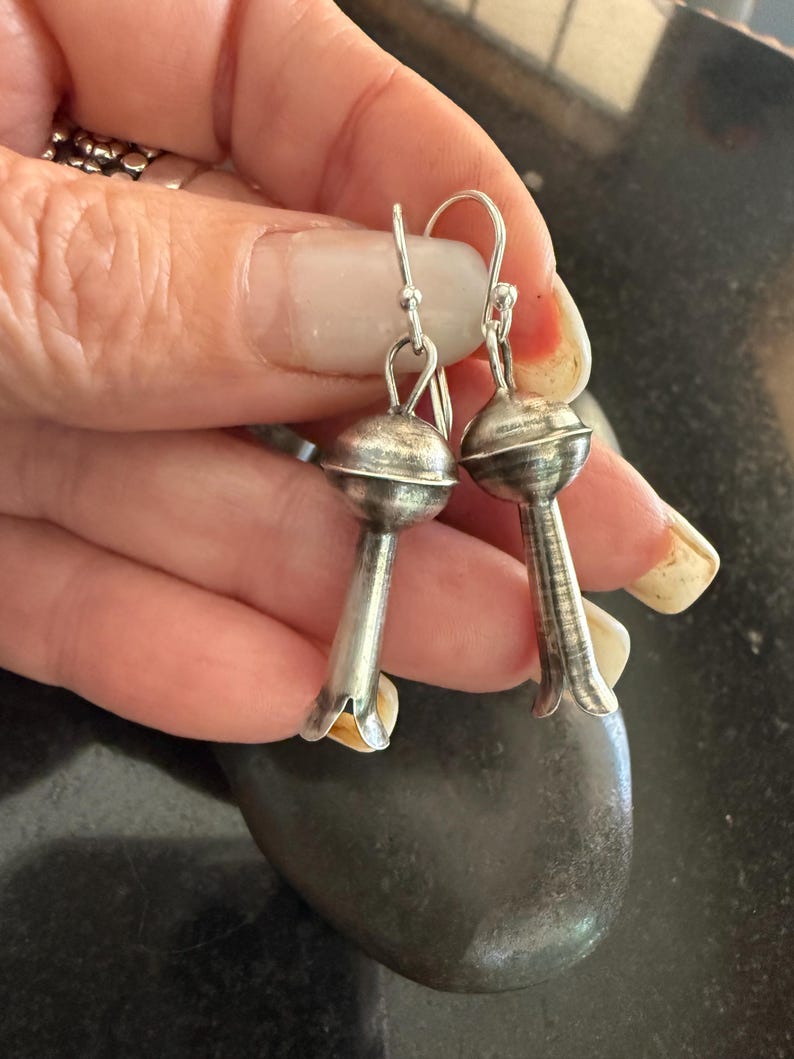 May include: A pair of silver earrings with a distinctive floral design. Each earring has a round top and a bell-shaped bottom. The earrings are suspended by a silver hook. The earrings are displayed on a dark surface.