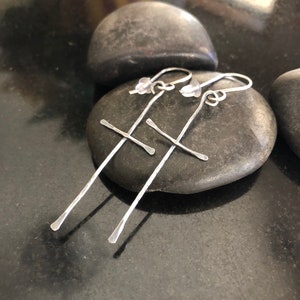 May include: A pair of silver cross earrings with a hammered finish. The earrings are hanging from a smooth gray stone.