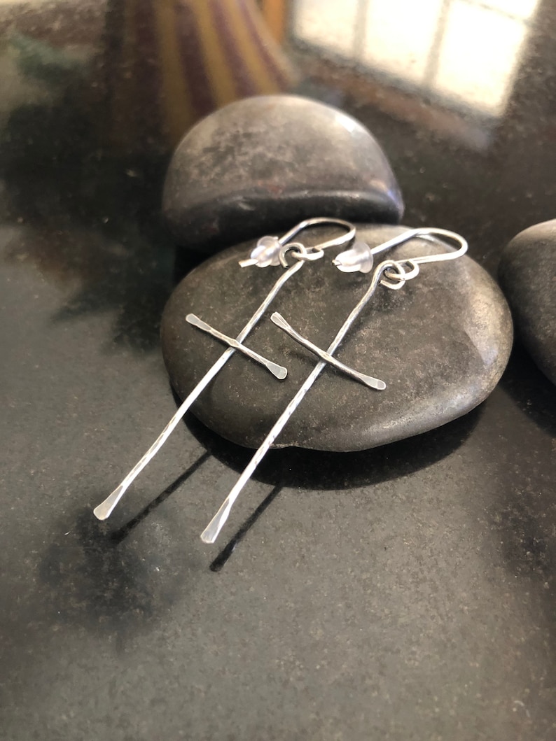 May include: A pair of silver cross earrings with a hammered finish. The earrings are hanging from a smooth gray stone.