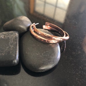 May include: A pair of hammered copper hoop earrings. The earrings have a textured surface and a warm, reddish-brown color. They are resting on a smooth, dark gray stone, with other stones in the background. The earrings are open at the bottom.
