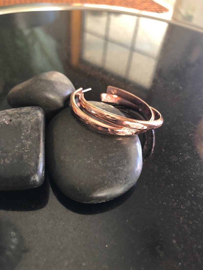 May include: A pair of hammered copper hoop earrings. The earrings have a textured surface and a warm, reddish-brown color. They are resting on a smooth, dark gray stone, with other stones in the background. The earrings are open at the bottom.