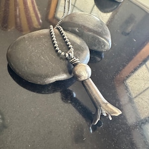 May include: A silver necklace with a unique pendant shaped like a flower. The pendant is made of metal and has a textured surface. The necklace is on a black background.