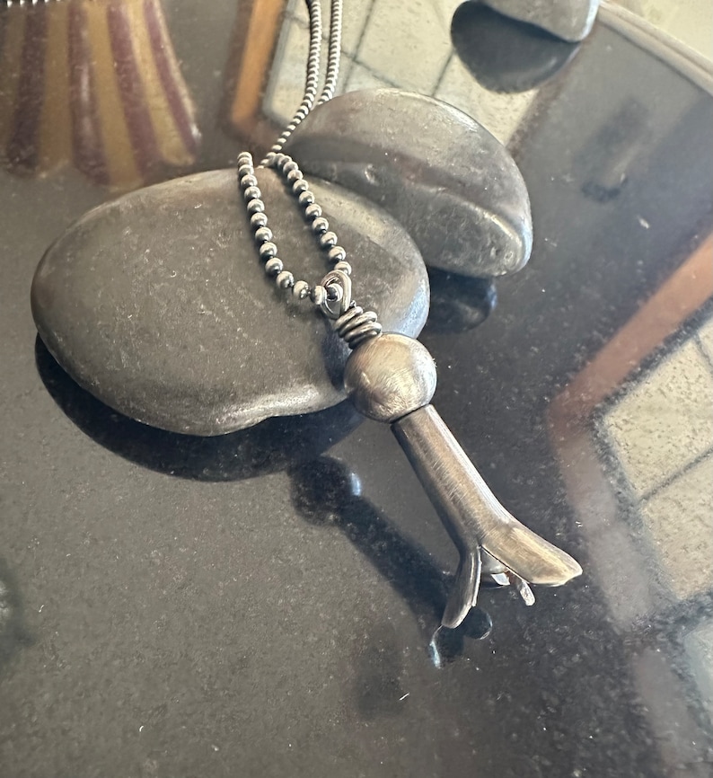 May include: A silver necklace with a unique pendant shaped like a flower. The pendant is made of metal and has a textured surface. The necklace is on a black background.