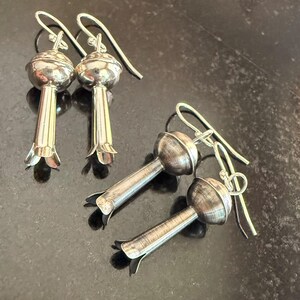 May include: Silver earrings with a floral design. Each earring has a rounded top and a long, slender stem with a flared, petal-like base. The ear wires are simple, curved hooks.