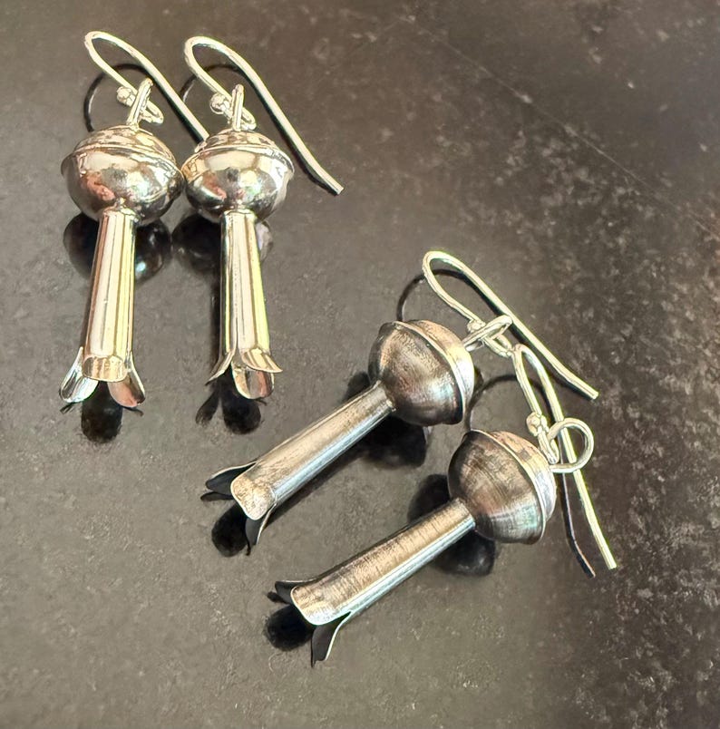 May include: Silver earrings with a floral design. Each earring has a rounded top and a long, slender stem with a flared, petal-like base. The ear wires are simple, curved hooks.