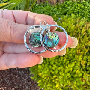 May include: Silver hoop earrings with a hammered finish and a round abalone shell pendant. The shell has iridescent green, blue, and purple colors.