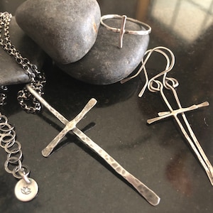 May include: A collection of handcrafted silver jewelry, including a necklace, ring, and earrings. The necklace features a chain with a pendant. The earrings and ring have a cross design. The jewelry is displayed on a dark surface with a natural stone.
