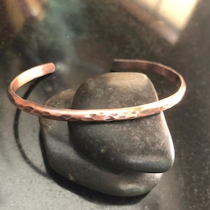 May include: A hammered copper cuff bracelet. The open-ended bracelet has a textured surface and is resting on a dark gray stone. The cuff is a simple, elegant design, suitable for everyday wear. The copper has a warm, reddish-brown hue.