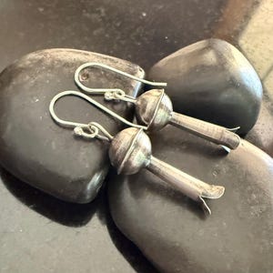 May include: A pair of silver-toned earrings with a unique, stylized floral design. Each earring features a rounded top and a slender, elongated stem with a flared, petal-like end. The earrings are displayed on dark, smooth stones.