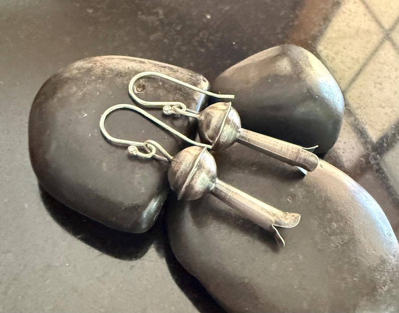 May include: Silver-coloured earrings with a distinctive floral design. Each earring has a rounded top and a slender stem with a flared, petal-like end. The earrings are presented on dark, smooth stones.