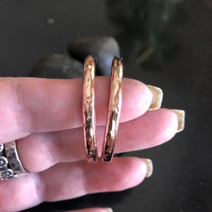 May include: A pair of hammered copper hoop earrings. The earrings have a textured surface and a warm, reddish-brown color. The hoops are held between fingers against a dark background.