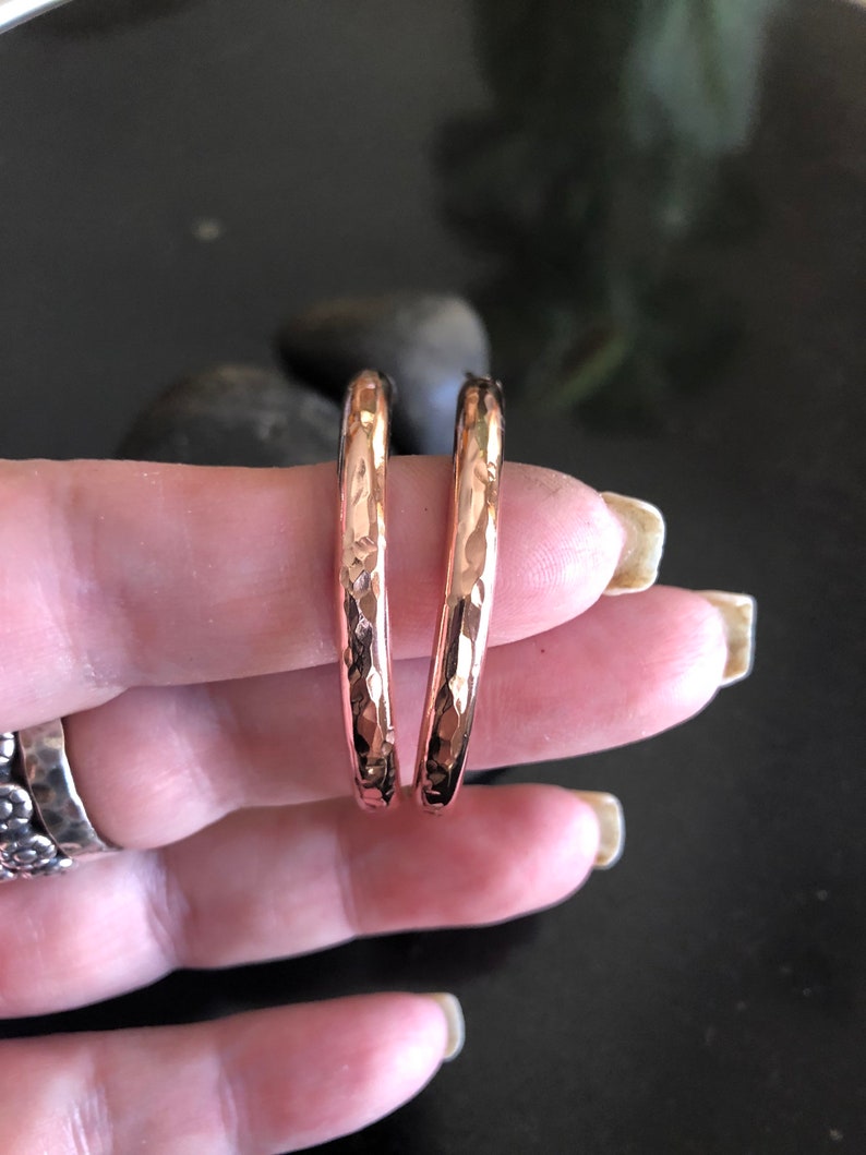 May include: A pair of hammered copper hoop earrings. The earrings have a textured surface and a warm, reddish-brown color. The hoops are held between fingers against a dark background.