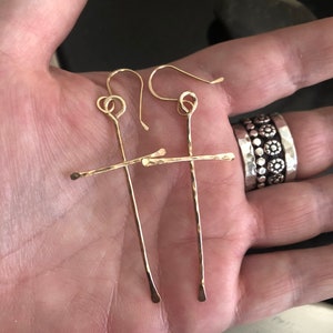 May include: A pair of gold-colored, handmade earrings. Each earring features a long, vertical bar crossed by a shorter horizontal bar, creating a cross shape. The earrings have a hammered texture and are displayed on a hand.