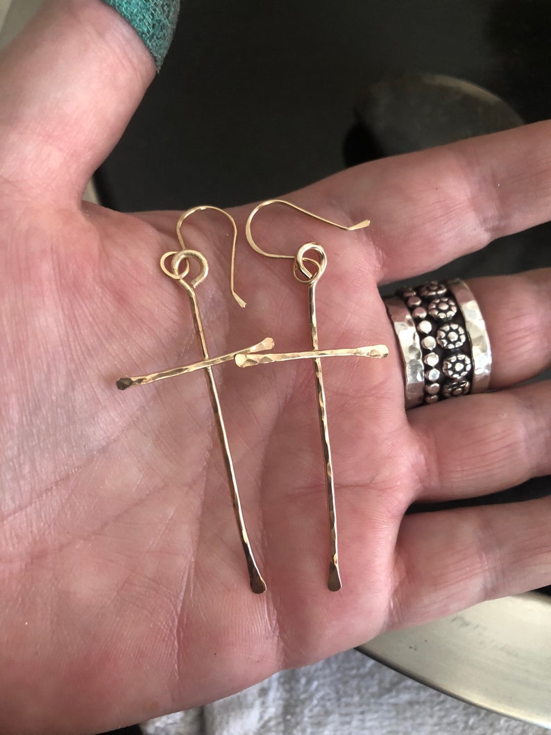 May include: A pair of gold-colored, handmade earrings. Each earring features a long, vertical bar crossed by a shorter horizontal bar, creating a cross shape. The earrings have a hammered texture and are displayed on a hand.