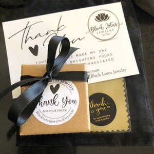May include: A small, square, brown jewelry box tied with a black satin ribbon. A white card with the text "Thank You" and a black heart is visible, along with a black drawstring bag. The box has a sticker that says "Thank You" and the Etsy shop's URL.