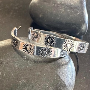 May include: A pair of silver hoop earrings with a sunburst design. The earrings are made of a shiny metal and feature a series of sunburst patterns around the hoops. The earrings are resting on a dark, textured surface.