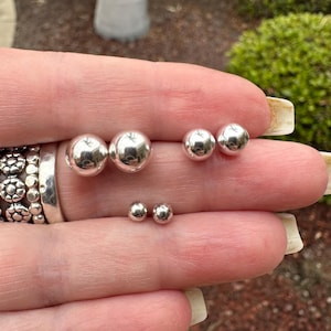 May include: A set of three pairs of silver ball stud earrings. The earrings are shown on a hand, with the largest pair on the index finger, the medium pair on the middle finger, and the smallest pair on the ring finger.