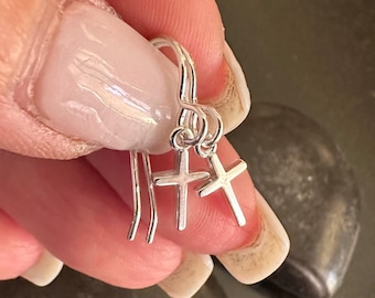 May include: Silver dangle earrings with small cross charms. The earrings are shown being held by a hand.
