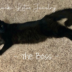 May include: A black cat with yellow eyes is lying on a carpet. The text "Black Lotus Jewelry" is written above the cat, and the text "- The Boss -" is written below the cat.